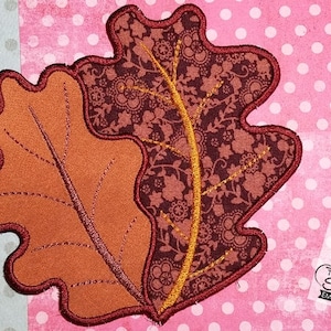 May include: Two brown felt oak leaves with a burgundy and gold floral pattern on one leaf. The leaves are stitched together and have a burgundy outline.