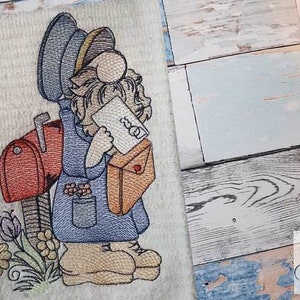Op de afbeelding: Borduurwerk van een kabouter die is verkleed als postbode, met een brief en een rode brievenbus. De kabouter draagt een blauwe hoed, een blauw jasje en bruine laarzen. Het ontwerp is omgeven door groen gebladerte en bloemen.