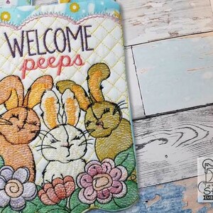May include: Embroidered Easter-themed quilted item with the words "WELCOME peeps" above three smiling bunnies and colorful flowers. The item has a light blue scalloped border and a diamond pattern.