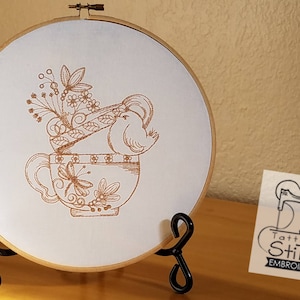 Może przedstawiać: Haftowany obręcz z brązową filiżanką do herbaty, na której brzegu siedzi ptak, i kwiatowymi akcentami. Obręcz stoi na czarnym stojaku. Obraz zawiera tekst "Tattered Stitch Embroideries".