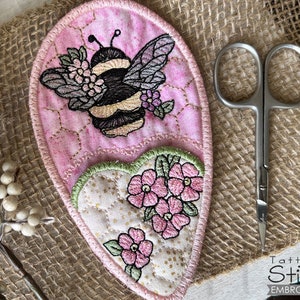 May include: A pink and green heart-shaped fabric applique with a bee and flowers embroidered on it. The bee is black and yellow with a pink flower on its back. The flowers are pink and white. The applique is on a burlap background.