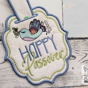 May include: A white fabric tag with a blue border and green stitching. The tag features a colorful bird with the text "Happy Passover" in green and blue lettering.
