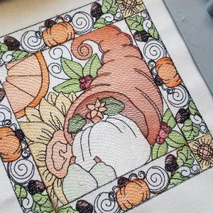 Könnte beinhalten: Stickerei mit einem Füllhorn, das überquillt mit herbstlichen Elementen wie Kürbissen, Sonnenblumen und Blättern. Das Design ist mit einem dekorativen Rand aus Spiralen und Eicheln umrahmt.