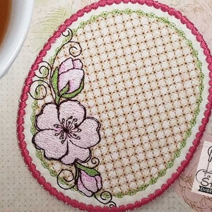 May include: A pink and green embroidered coaster with a floral design. The coaster is oval shaped and has a scalloped edge.