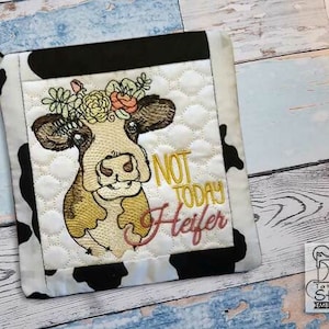 May include: A black and white cow with a flower crown is embroidered on a white pot holder. The pot holder has a black border and a black and white cow print background. The text "Not Today Heifer" is embroidered in yellow and pink.