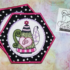 May include: Two hexagon-shaped fabric coasters with a pink border and black and white polka dots. The coasters feature an embroidered design of a teapot with a heart and swirls in pink, green, and purple.