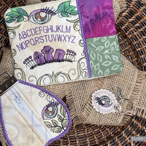 May include: A set of three embroidered pouches with a spooky theme. The largest pouch features an eye, a row of letters, and a row of flowers. The medium pouch is a teardrop shape with an eye and floral designs. The smallest pouch is a round charm with an eye. All three pouches are made of beige fabric with purple stitching.