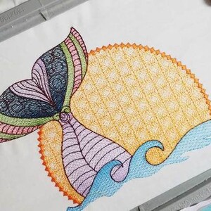 Puede incluir: Diseño bordado de colores de una cola de ballena con un sol y olas. La cola está rayada de rosa, verde, azul y morado. El sol es amarillo con un patrón texturizado. Las olas son azules.