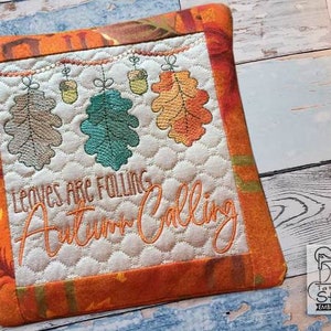 May include: A white quilted pot holder with an orange border. The centre features an embroidered design of leaves falling from a branch with the text "Leaves are falling Autumn Calling".