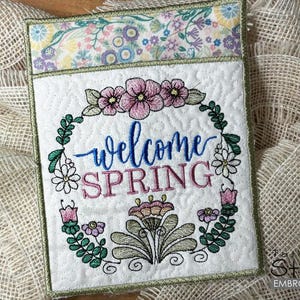 Op de afbeelding: Een witte stoffen deurhanger met een groene bloemenkrans en de tekst "Welcome Spring". De krans is versierd met roze bloemen en groene bladeren.