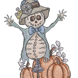 Puede incluir: Diseño bordado de un espantapájaros con rostro cosido, que lleva un sombrero con una flor y una pajarita azul. El espantapájaros se alza sobre tres calabazas naranjas, con detalles de hojas. El diseño es en tonos beige, azul y naranja.