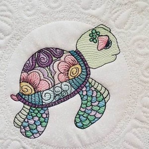 Può includere: Una tartaruga ricamata colorata con un motivo floreale sul guscio e un motivo verde e blu sulle zampe. La tartaruga è su uno sfondo bianco.