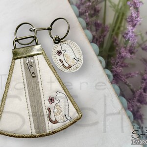 May include: A white fabric zippered pouch with a brown trim and a small embroidered cat design. The pouch is attached to a key ring with a small round charm featuring a similar cat design.