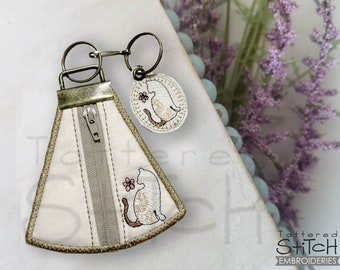 Può includere: Una pochette in tessuto bianco con cerniera, bordatura marrone e un piccolo disegno di gatto ricamato. La pochette è attaccata a un portachiavi con un piccolo ciondolo rotondo con un disegno di gatto simile.