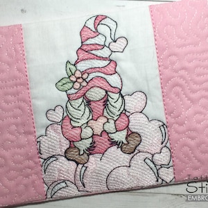 Puede incluir: Diseño bordado de un gnomo rosa y blanco con un gorro rojo y blanco, rodeado de corazones rosas y nubes. El diseño está sobre una tela acolchada rosa con un borde blanco.
