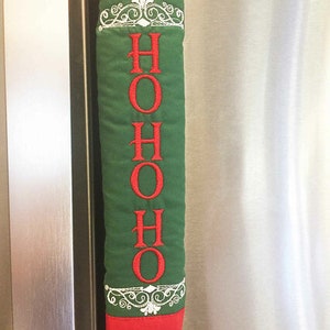 Op de afbeelding: Een groene stoffen deurgreephoes met rode bies en witte borduurwerk. Het borduurwerk zegt "HO HO HO" met een decoratief ontwerp erboven en eronder.