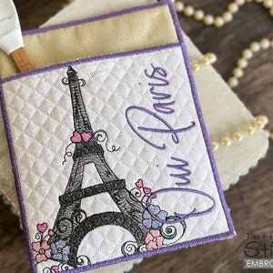 May include: A white quilted pot holder with a purple border features an embroidered design of the Eiffel Tower with pink flowers and the text "Oui Paris".