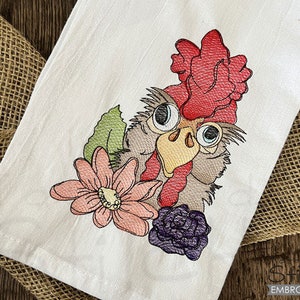 May include: White kitchen towel featuring an embroidered rooster with a red comb, gray feathers, and large, expressive eyes. The design includes a pink flower, green leaf, and purple flower. The towel is folded on a burlap surface.