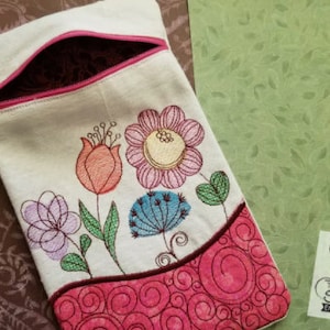 May include: A white zippered pouch with a pink zipper and a floral embroidery design. The embroidery features pink, blue, green, and orange flowers. The pouch has a pink and white swirl design at the bottom.
