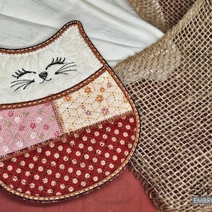 Puede incluir: Un posavasos acolchado con forma de gato, con una cara color crema, ojos, nariz y bigotes bordados. El cuerpo es un patchwork de telas florales y de lunares en rosa, crema y rojo. El posavasos está bordeado con costuras marrones.