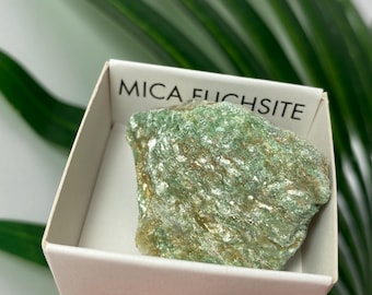 May include: A rough, green Mica Fuchsite stone rests inside a white box. The stone has a textured surface with flecks of white and brown. The box has the words "MICA FUCHSITE" printed on the top.
