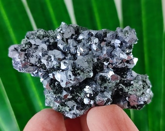 May include: A cluster of metallic gray mineral crystals with a reflective surface. The crystals are clustered together, creating a textured appearance. The mineral is held against a green background.