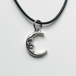 以下が含まれることがあります： 黒い紐のシルバー三日月ペンダントネックレス。三日月チャームには詳細な顔が描かれています。ネックレスは白い背景に表示されています。紐の長さは約40cmです。