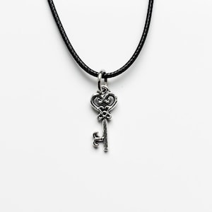 May include: A silver key pendant on a black cord necklace. The key features an elaborate design. The background is a simple white.