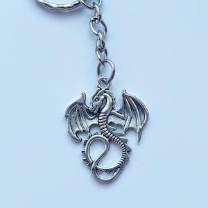May include: A silver dragon keyring. The dragon pendant features detailed wings and a coiled body. The keyring is attached to a silver chain and ring. The background is a plain, light colour.