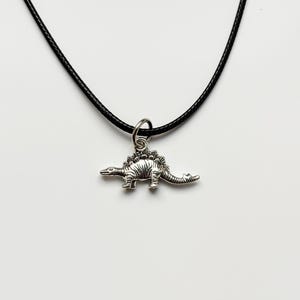 Pode incluir: Colar com pingente de dinossauro prateado. O pingente é uma representação detalhada de um Estegossauro. O colar tem um cordão preto. O fundo é branco.