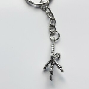 May include: A silver-colored keychain featuring a detailed bird claw charm. The claw is attached to a chain and a larger ring. The background is a plain white surface. The keychain is a unique accessory.