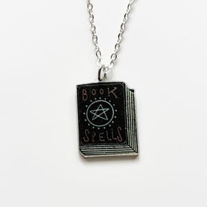 Peut inclure: Un collier à chaîne argentée avec un pendentif noir en forme de livre. Le pendentif porte les mots "BOOK SPELLS" et un symbole d'étoile. Le collier est présenté sur un fond blanc.