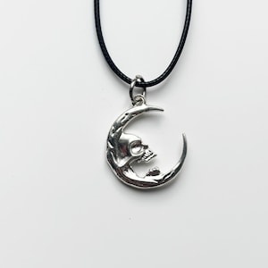 May include: Silver-toned pendant necklace featuring a crescent moon design with a skull face. The pendant hangs from a black cord. The necklace is displayed against a white background.