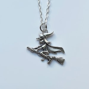 Pode incluir: Colar com pingente prateado de uma bruxa numa vassoura. A bruxa usa chapéu pontudo e capa. A corrente do colar é fina, sobre fundo branco.