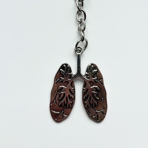 Puede incluir: Un llavero plateado con un colgante en forma de pulmones. El colgante presenta detalles anatómicos intrincados y está unido a una cadena y un anillo. El texto "Pissy Prints" es visible en la parte inferior.