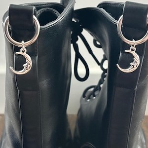 以下が含まれることがあります： 黒い革のコンバットブーツのペア。上部のループから銀色の三日月チャームがぶら下がっています。ブーツには白いステッチと黒いレースが付いています。