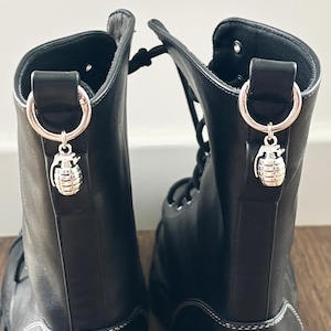 Può includere: Stivali da combattimento in pelle nera con ciondoli a forma di granata color argento. Gli stivali hanno cuciture bianche e un design stringato. Gli stivali sono alti circa 25 cm.
