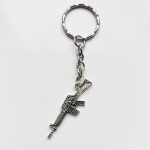 May include: Silver-tone metal keychain with a rifle-shaped charm. The keychain has a ring and chain. The text "Pissy Prints" is printed at the bottom of the image.
