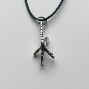 Puede incluir: Collar con colgante plateado en forma de pata de pollo detallada. El colgante, con diseño de garra, cuelga de un cordón negro. El collar presenta un amuleto realista de pata de ave, mostrando detalles de garras y escamas.