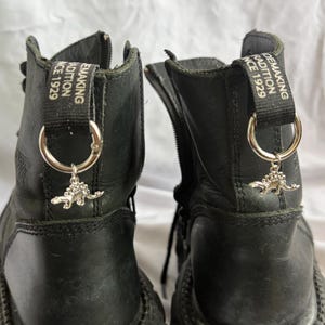 Könnte beinhalten: Ein Paar schwarze Lederstiefel mit einem silberfarbenen Dinosaurier-Anhänger an einem Metallring an der Fersenzuglasche. Die schwarze Stofflasche zeigt den weißen Text "SHOEMAKING TRADITION SINCE 1929" und hat einen seitlichen Reißverschluss.