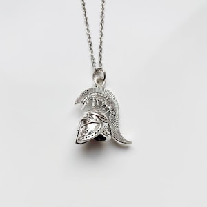 May include: Silver-toned pendant necklace featuring a detailed Spartan helmet design. The pendant hangs from a delicate chain, showcasing the helmet's intricate details. The necklace is displayed against a plain white background.