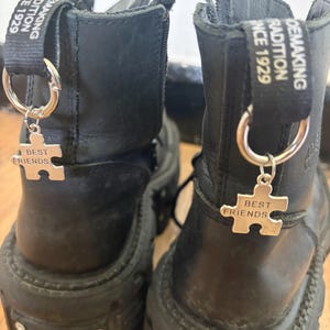 Può includere: Un paio di stivali neri in pelle con suole spesse. Ogni stivale ha un ciondolo d'argento a forma di pezzo di puzzle con la scritta "BEST FRIENDS". Gli stivali hanno un passante in tessuto con la scritta "TRADITION SINCE 1929".