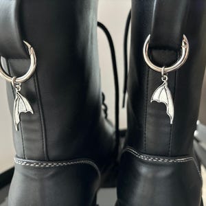 May include: A pair of black leather boots with silver wing charms attached to the laces. The boots have a white stitching detail around the top of the boot.