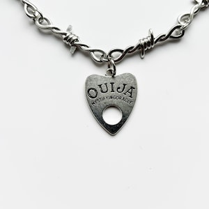 May include: Silver-coloured necklace with a barbed wire design. A heart-shaped pendant with the word "OUJIA" and "MYSTIFYINGORACLE" is suspended from the necklace. The necklace is shown against a white background.