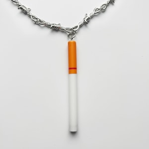 May include: A silver-tone necklace with a barbed wire chain. The pendant is a cigarette with an orange filter and a red band. The cigarette is white and cylindrical in shape.
