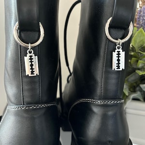 Può includere: Stivali in pelle nera con suola platform massiccia e un ciondolo a forma di lama da barba argentata su ogni stivale.