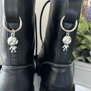 May include: A pair of black leather boots with silver charms shaped like aliens hanging from the laces. The boots have a chunky platform sole and white stitching around the top.
