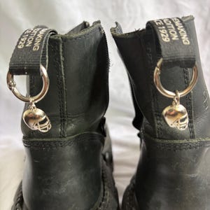 Può includere: Un paio di stivali in pelle nera con una linguetta nera sul retro di ogni tallone con il testo bianco "MAKING TRADITION SINCE 1929". Ogni linguetta include un anello in metallo argentato con un piccolo ciondolo a forma di casco da football.