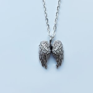 May include: A silver-colored necklace featuring a pendant shaped like angel wings. The wings are detailed with textured feather patterns and are suspended from a delicate chain. The necklace is displayed against a light blue background.