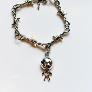 May include: A silver-toned metal bracelet featuring a chain designed to resemble barbed wire. The bracelet includes a central charm shaped like a stylized alien figure with a large head and smooth, reflective surface, finished with a lobster clasp.
